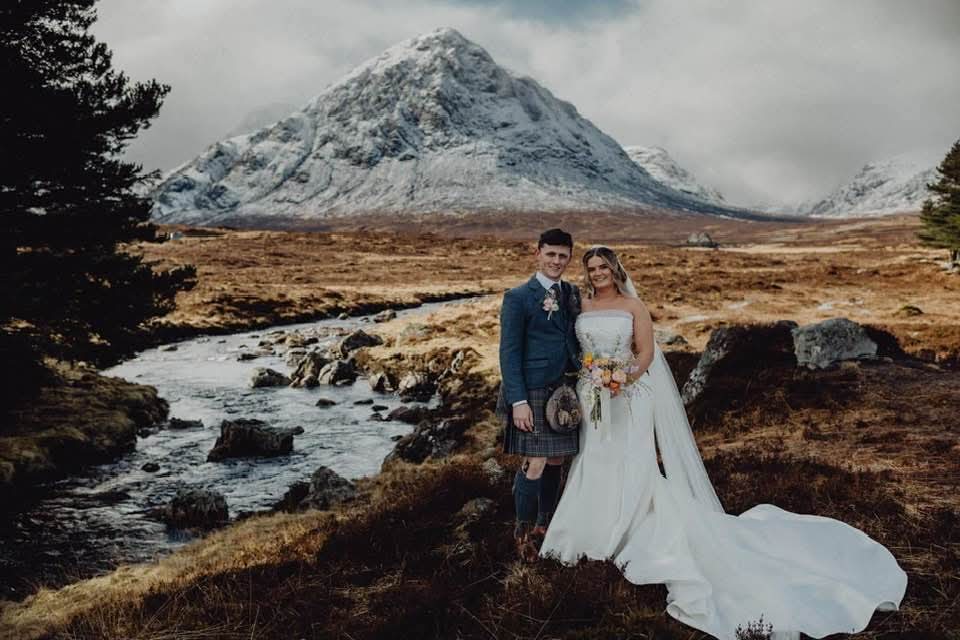 Bride and groom walking hand in hand in a scenic Highland landscape, with a river and snow-capped mountain in the background, the groom wearing a traditional kilt and the bride holding a colorful bouquet.