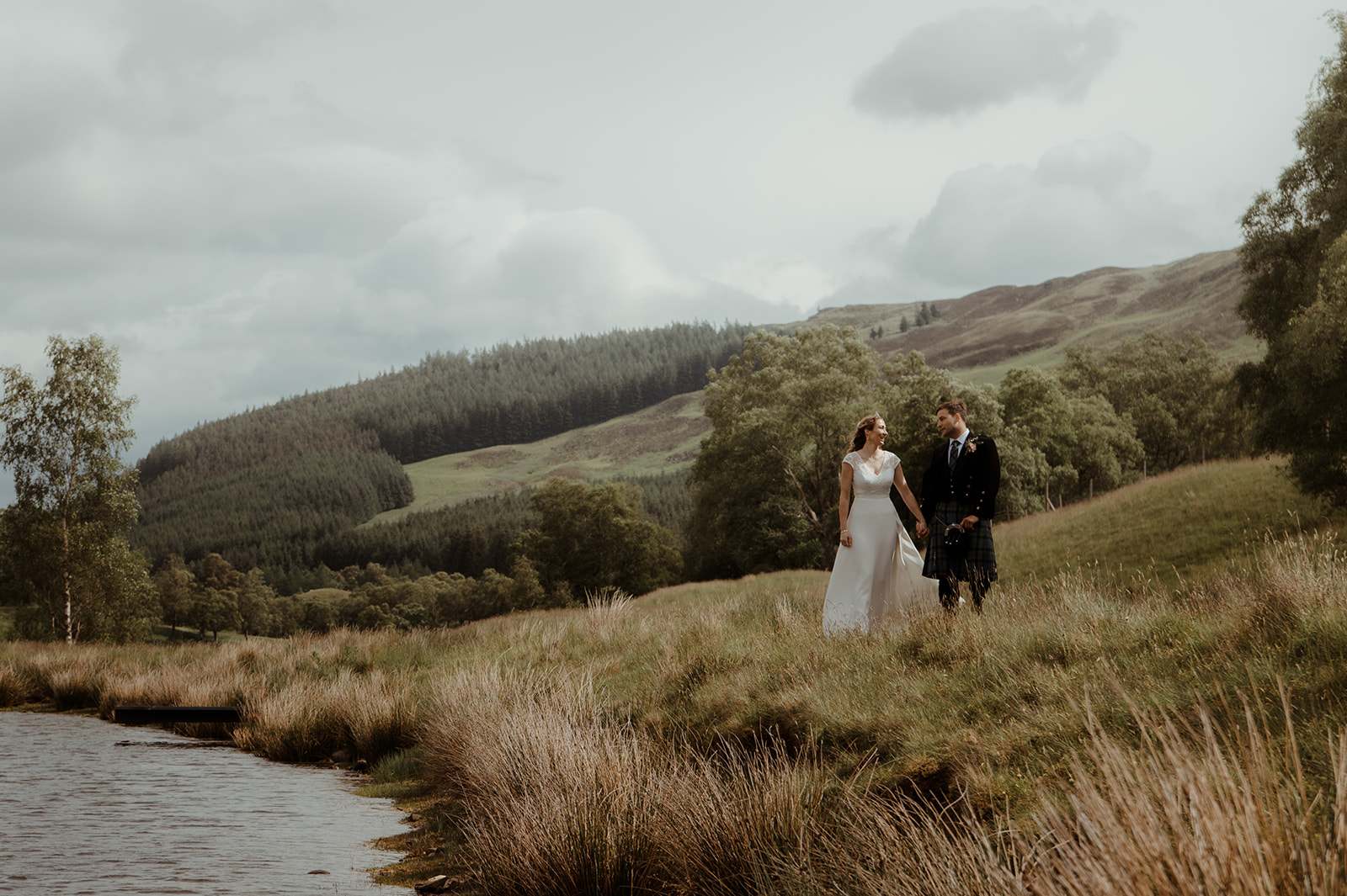bride and groom walking hand in hand through grassy countryside near a lake, surrounded by rolling hills and trees under a cloudy sky.