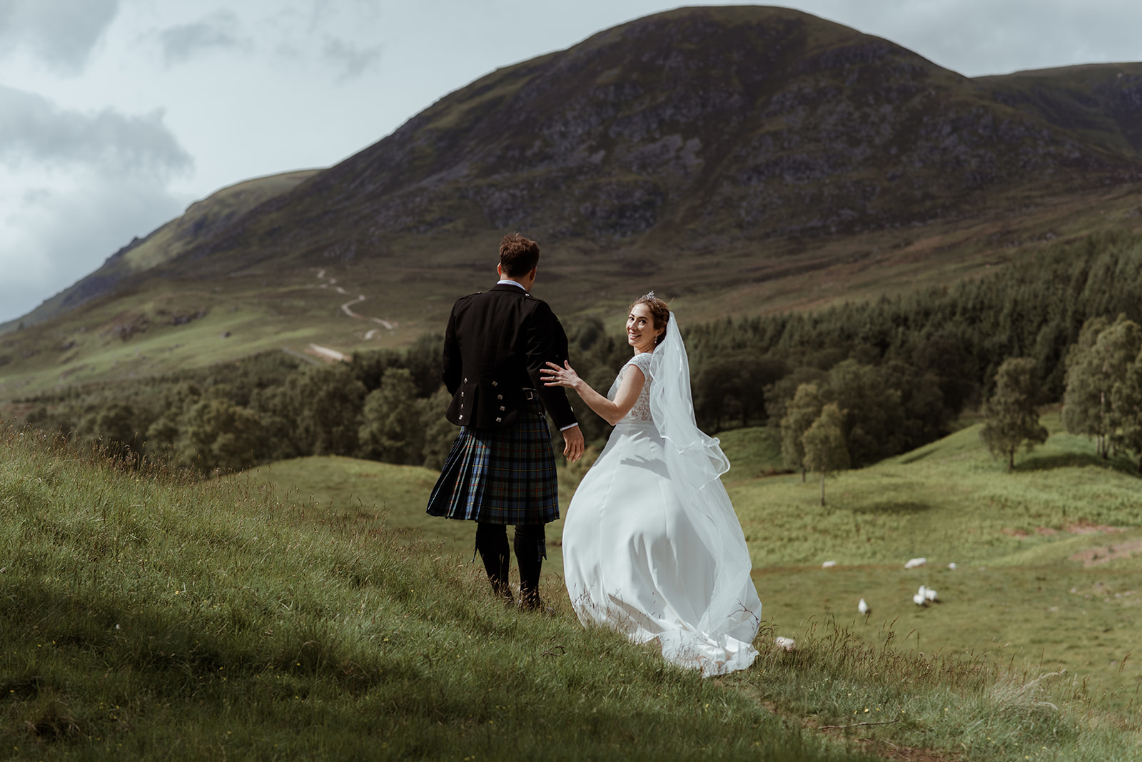 bride in white gown and groom in traditional kilt walking across a grassy hillside with mountain backdrop, scenic countryside wedding portrait.