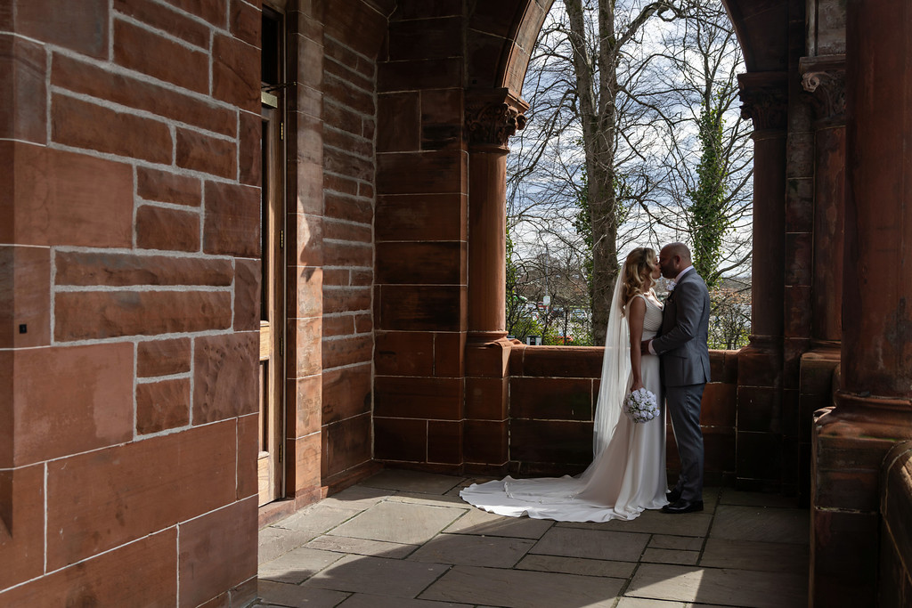 Bride and groom sharing a kiss in a stone archway, with warm red brick walls, natural light, and trees visible through the opening.