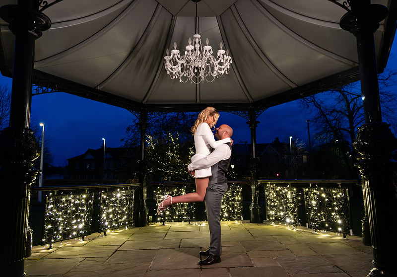 Bride and groom embracing under a gazebo at night, with the groom lifting the bride as they kiss beneath a chandelier, surrounded by soft string lights.