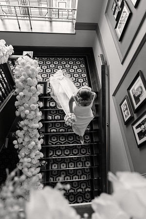 Black-and-white overhead view of a bride walking down a patterned staircase, holding her dress, with floral decorations along the railing.