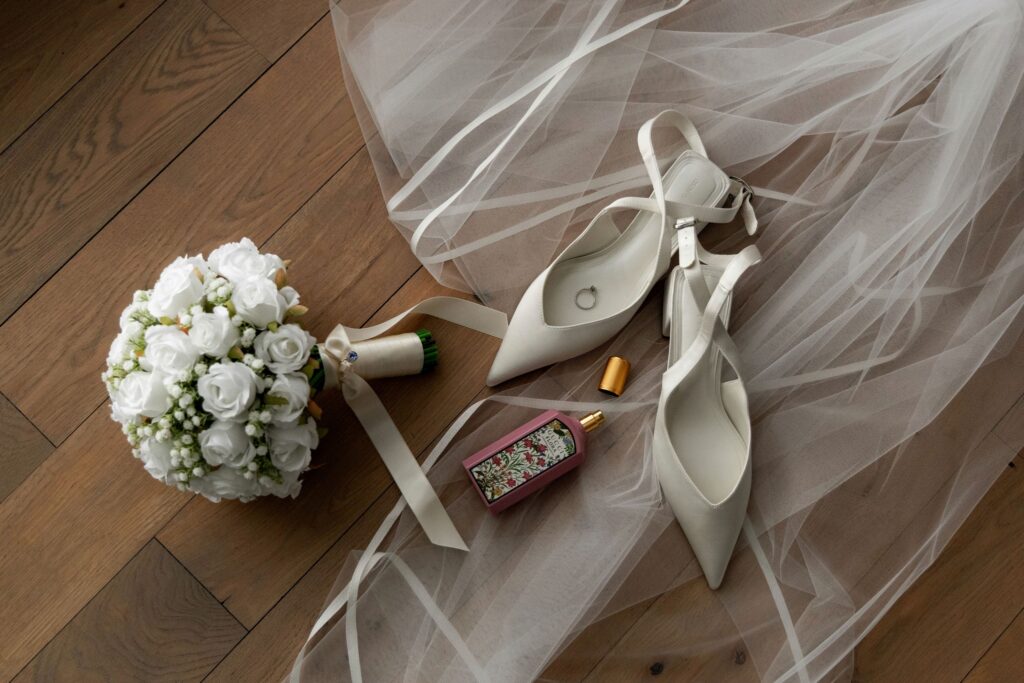 Flat lay of bridal details on a wooden floor, including white heels with a ring, a bouquet of white roses, a perfume bottle, and soft tulle fabric.
