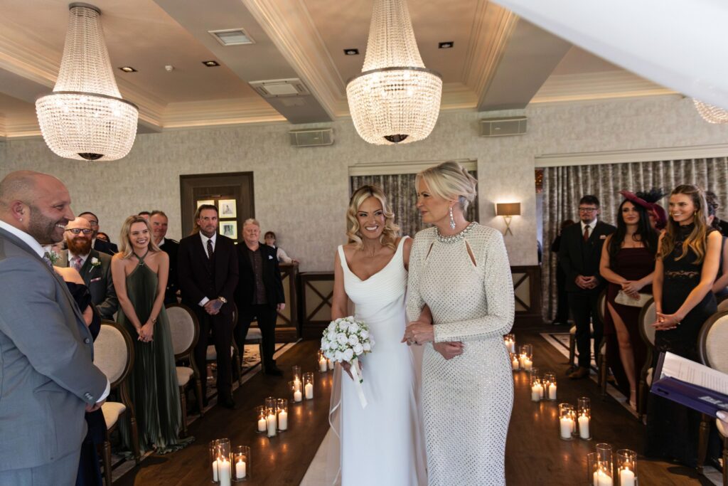 Bride walking down the aisle with an older woman, smiling and holding a bouquet, surrounded by guests and candlelit decor in an elegant indoor wedding ceremony.