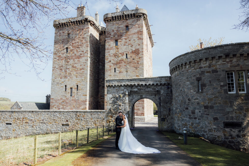 Bride and groom embracing in front of the historic entrance of Borthwick Castle during an elegant outdoor wedding portrait.