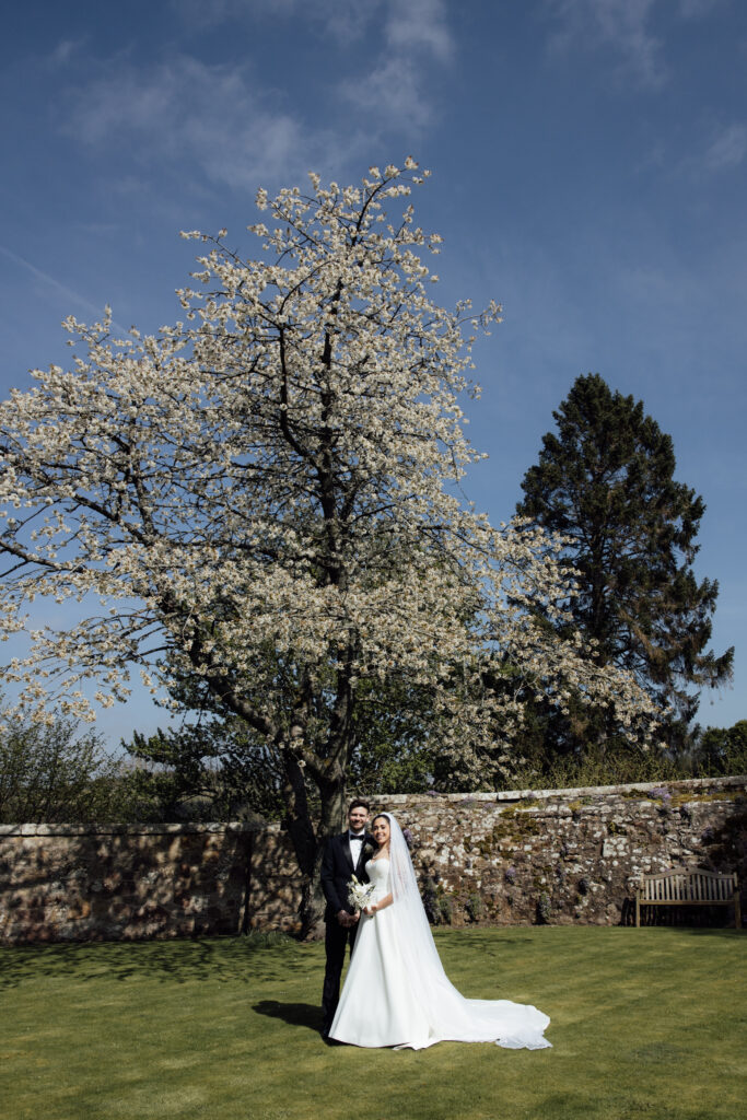 Bride and groom posing beneath a large blooming tree in the gardens of Borthwick Castle on a bright spring wedding day.