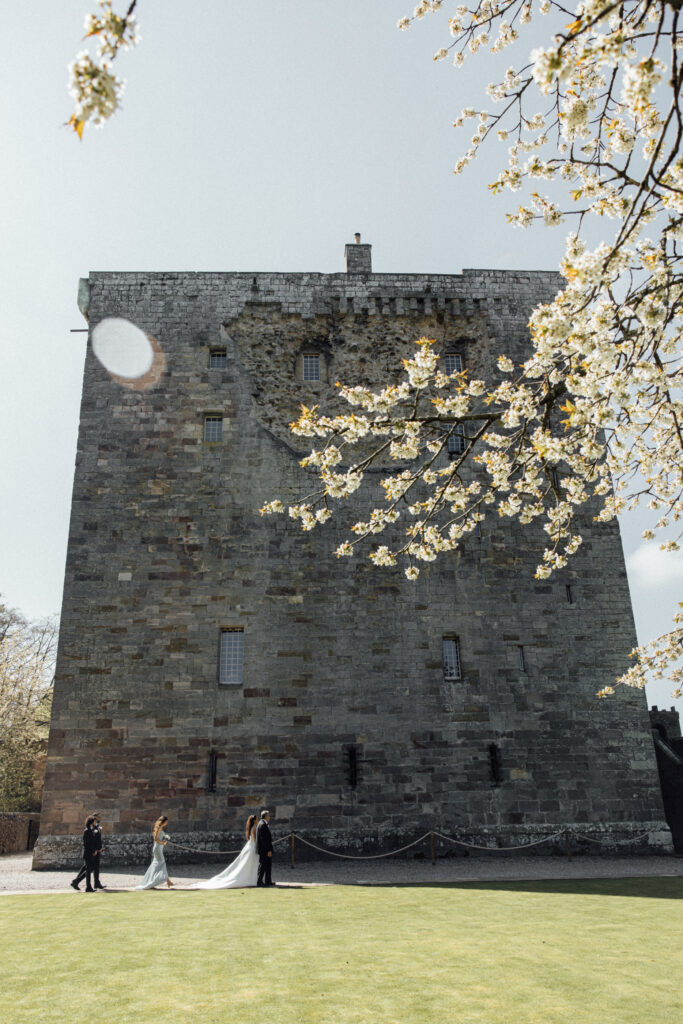 Wide view of Borthwick Castle with a bride and groom walking across the grounds beneath blooming spring trees on a sunny wedding day.