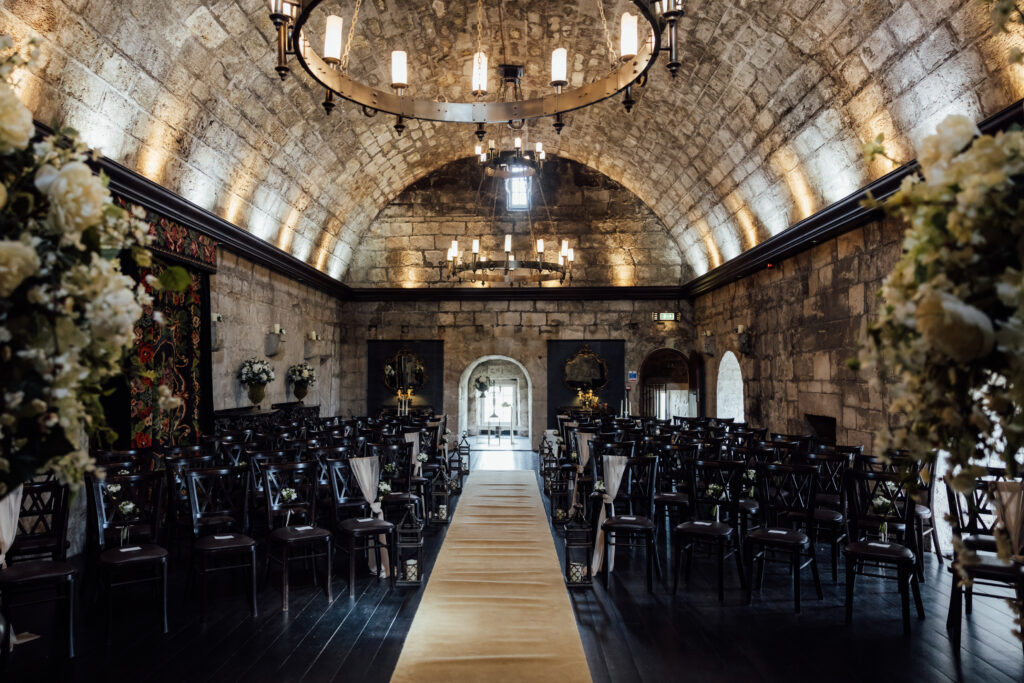 Elegant wedding ceremony setup inside Borthwick Castle featuring stone arch ceilings, candle-style chandeliers, floral arrangements, and a central aisle lined with dark wooden chairs.
