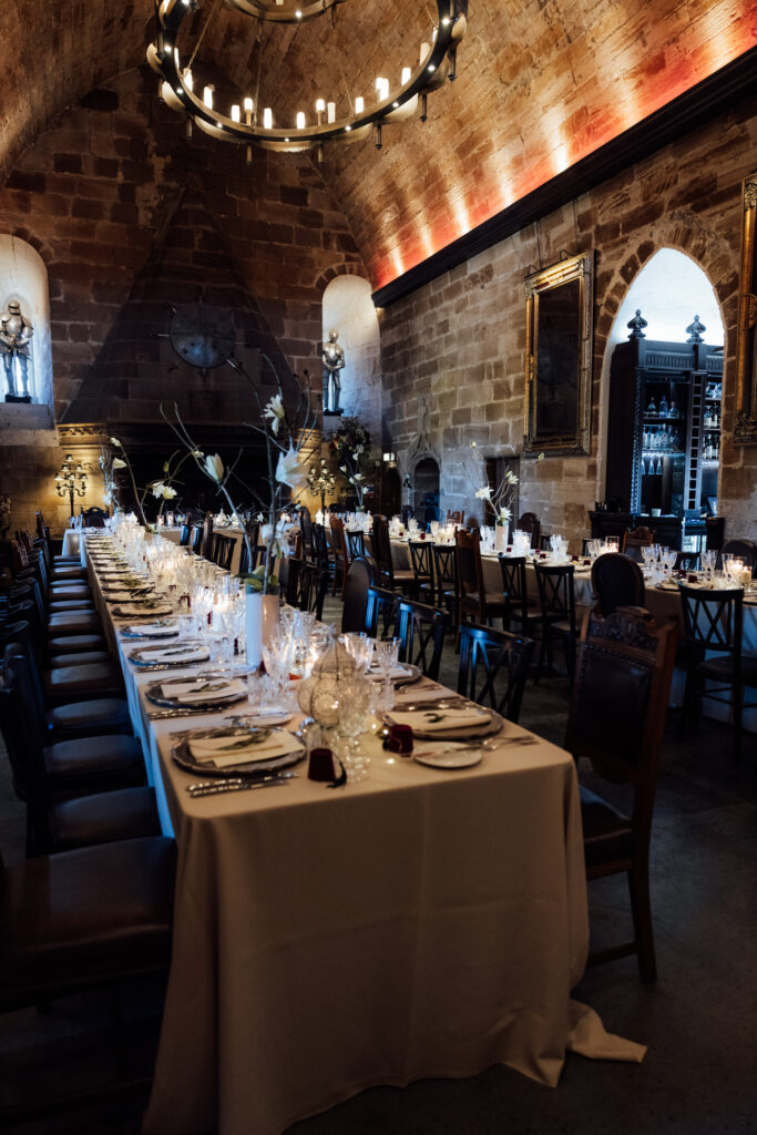 Elegant wedding reception setup inside Borthwick Castle featuring long candlelit dining tables, vaulted stone ceilings, chandeliers, and medieval-inspired décor.