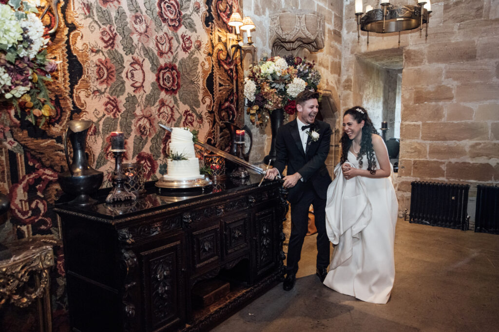 Bride and groom laughing together while cutting their wedding cake with a ceremonial sword inside the historic halls of Borthwick Castle.