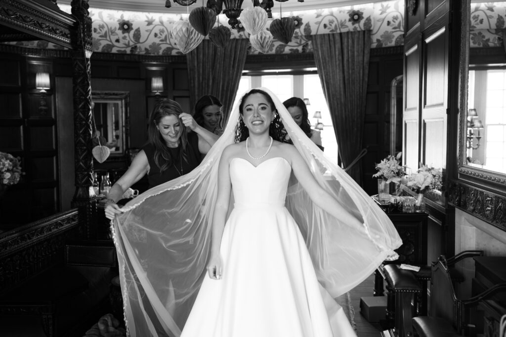 Black-and-white portrait of a smiling bride in a wedding gown as bridesmaids help arrange her veil inside an elegant room at Borthwick Castle.