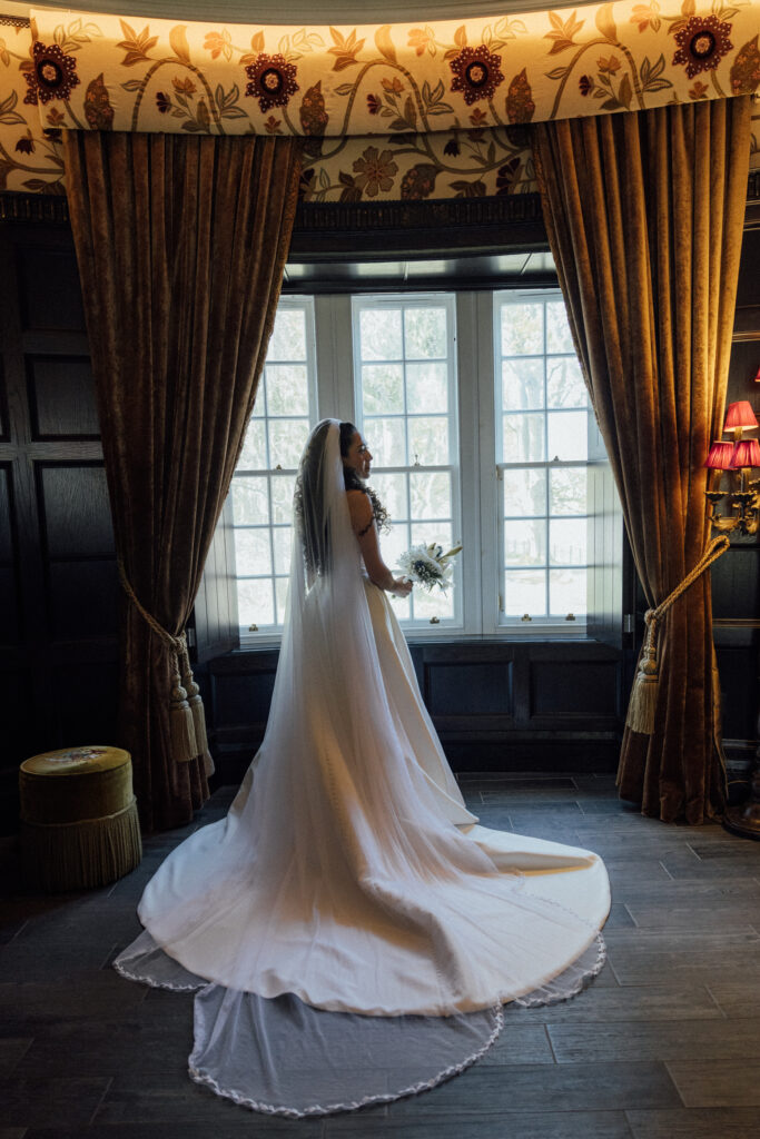 Bride in a flowing wedding gown standing by tall windows inside Borthwick Castle, surrounded by elegant floral wallpaper and rich velvet curtains.