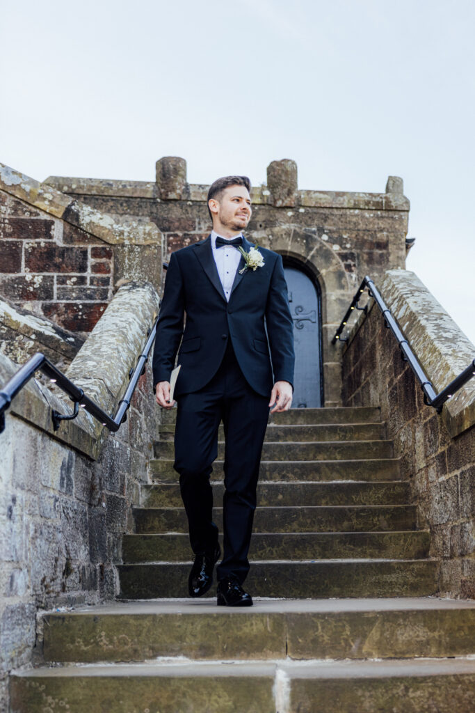 Groom in a black tuxedo walking down the historic stone steps of Borthwick Castle during an elegant outdoor wedding portrait.