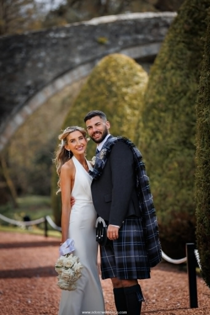 Bride and groom posing together beneath the historic Brig o’ Doon bridge in Alloway, with the groom wearing a traditional Scottish kilt and the bride holding a white bouquet during their romantic outdoor wedding photoshoot.