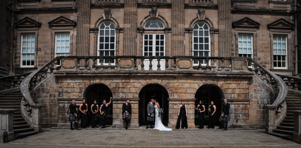 Bride and groom sharing a kiss beneath the grand stone archways of Sorn Castle, surrounded by their wedding party in coordinated attire, with the castle's impressive Georgian façade and sweeping staircases creating a stunning Ayrshire wedding backdrop.