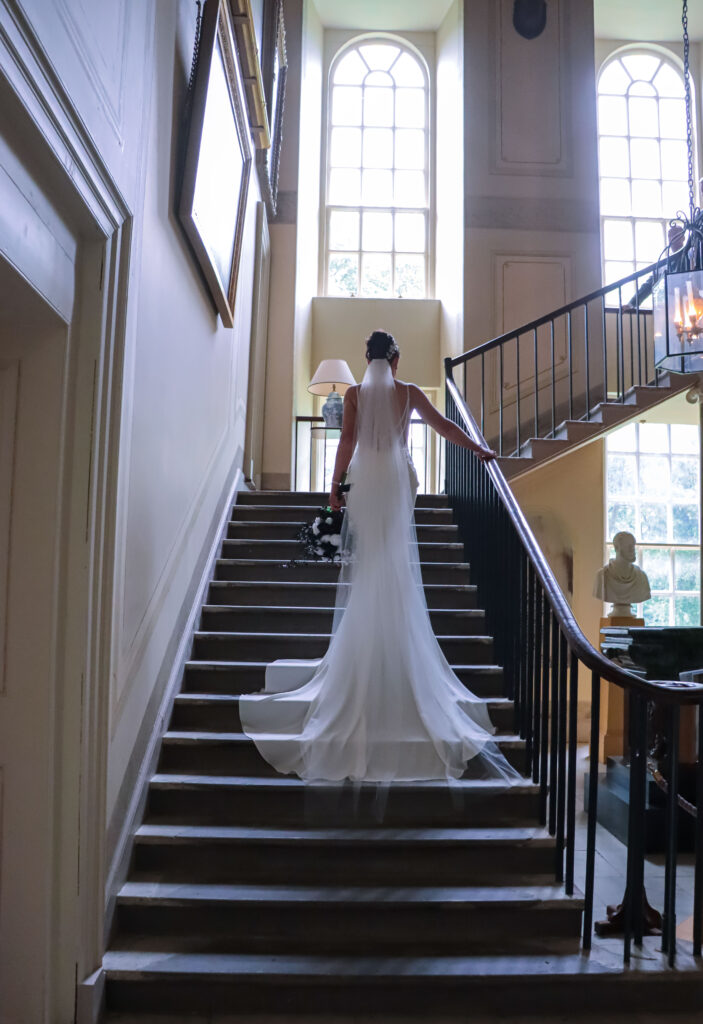Bride walking up the grand staircase inside Sorn Castle, holding her bouquet as natural light streams through tall arched windows, creating an elegant wedding portrait.