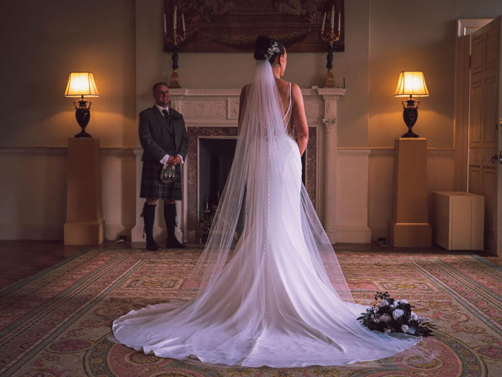 Bride standing in her elegant wedding gown and long veil inside Sorn Castle as the groom smiles during their emotional first look, with a grand fireplace and bouquet completing the romantic scene.