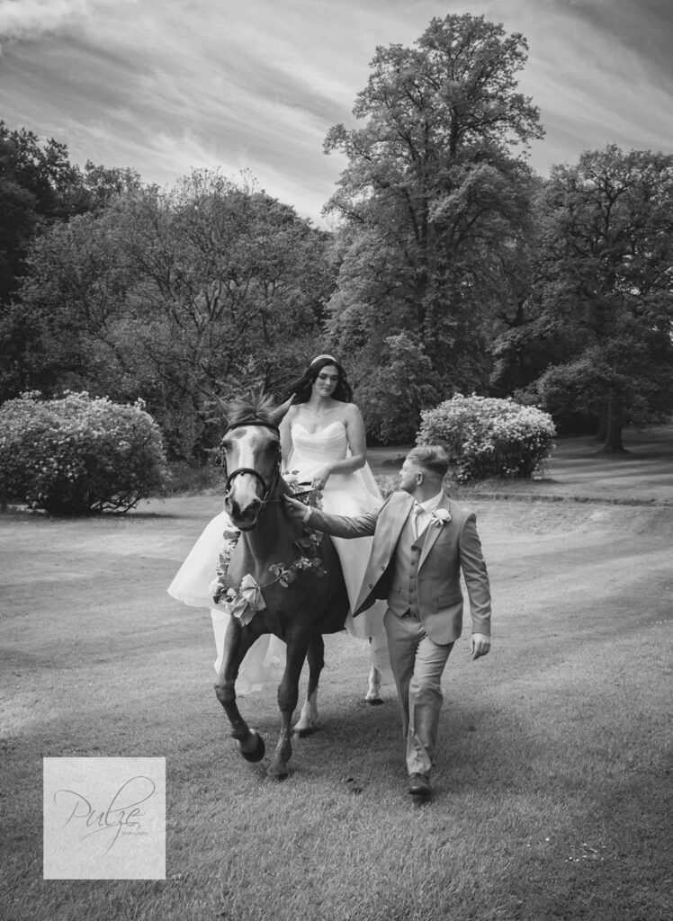 Black-and-white wedding portrait of a groom leading a flower-adorned horse while the bride rides through a scenic garden surrounded by mature trees.