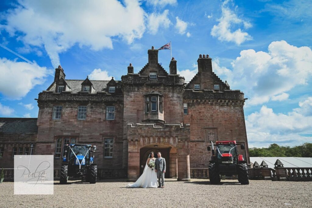 Bride and groom standing in front of a historic castle with blue and red tractors on either side under a bright blue sky, creating a unique countryside wedding portrait.