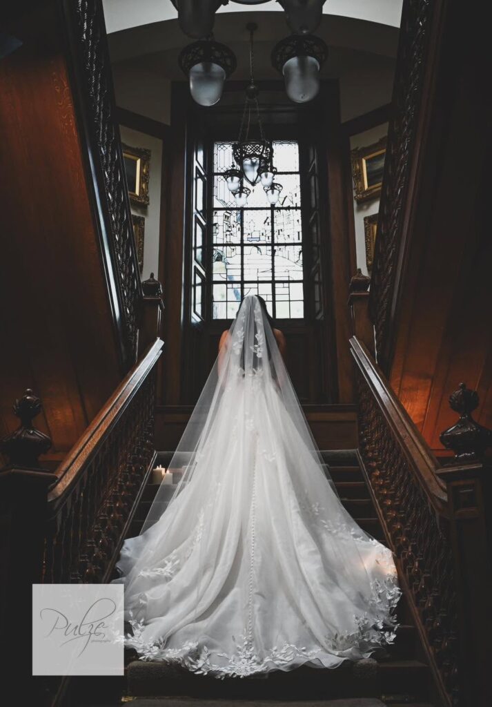 Bride in a flowing wedding gown and embroidered veil standing on a grand wooden staircase beneath a stained-glass window, creating an elegant bridal portrait.