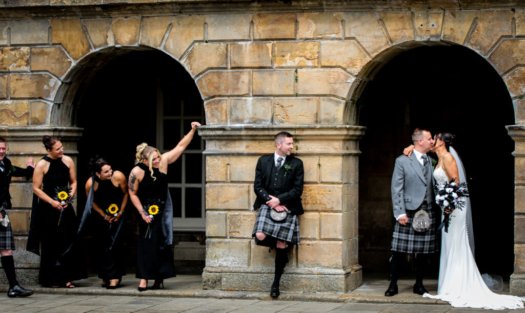 Bride and groom sharing a kiss beneath the stone archways of Sorn Castle while their wedding party playfully peeks around the columns, creating a fun and candid Ayrshire wedding portrait.