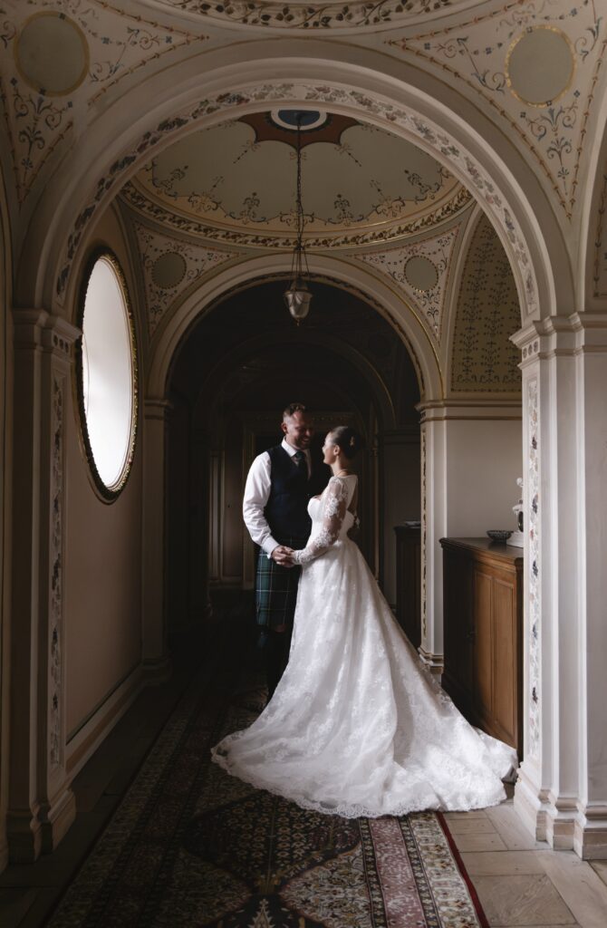 Bride and groom share a romantic moment beneath elegant arched ceilings at Dumfries House, with the bride wearing a lace wedding dress and the groom in a traditional Scottish kilt, beautifully framed by the venue's ornate historic interior.