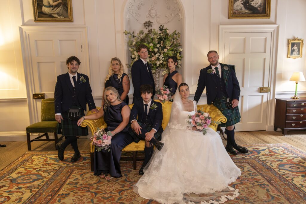 Bride and groom pose with their wedding party in an elegant reception room, with the bride seated in a lace wedding gown holding a pink and white bouquet and the groom wearing a traditional Scottish kilt. Bridesmaids and groomsmen in navy attire are arranged around a yellow sofa beneath a large floral display, creating a formal group wedding portrait.