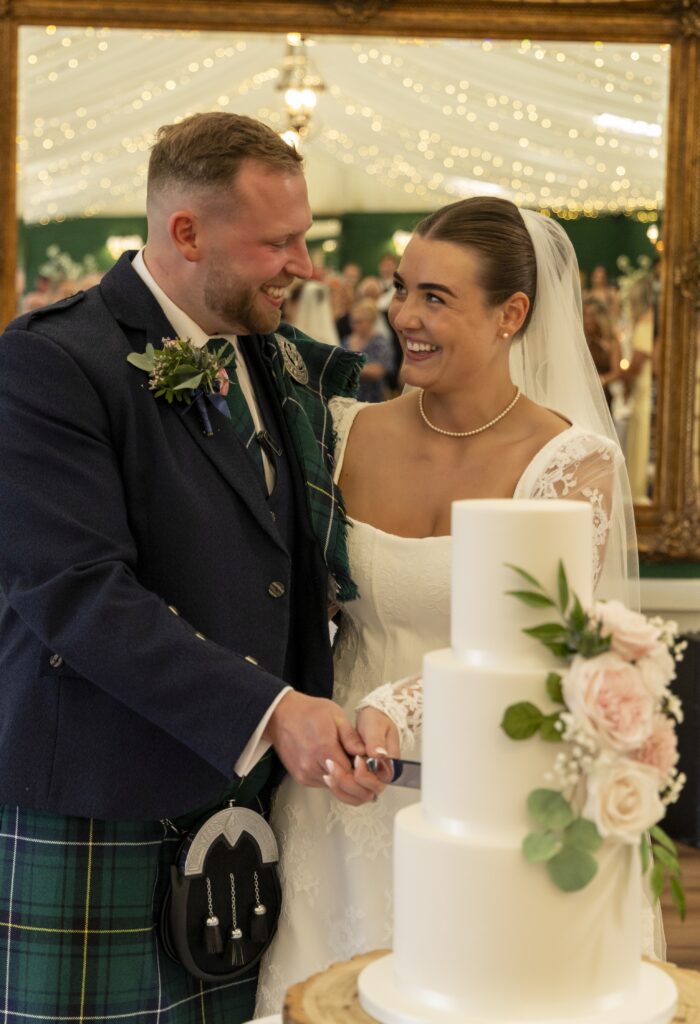 Bride and groom smiling at each other as they cut a three-tier white wedding cake decorated with blush roses and greenery. The groom wears a traditional Scottish kilt, while the bride wears a lace wedding dress and veil, surrounded by warm fairy lights at their wedding reception.