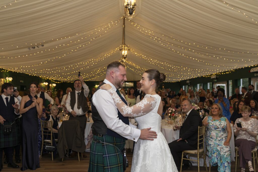 Bride and groom share their first dance beneath twinkling fairy lights at their wedding reception, smiling as they embrace on the dance floor. The bride wears a lace wedding dress, while the groom wears a traditional Scottish kilt, surrounded by family and friends watching the special moment inside a beautifully decorated marquee.