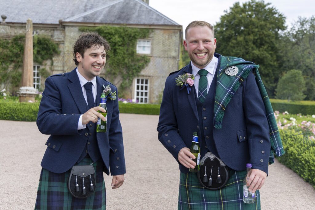 Groom and groomsman wearing traditional Scottish kilts smile and chat while holding drinks outside a historic country house. Surrounded by formal gardens, the relaxed candid wedding portrait captures the pair enjoying the celebration before the reception.