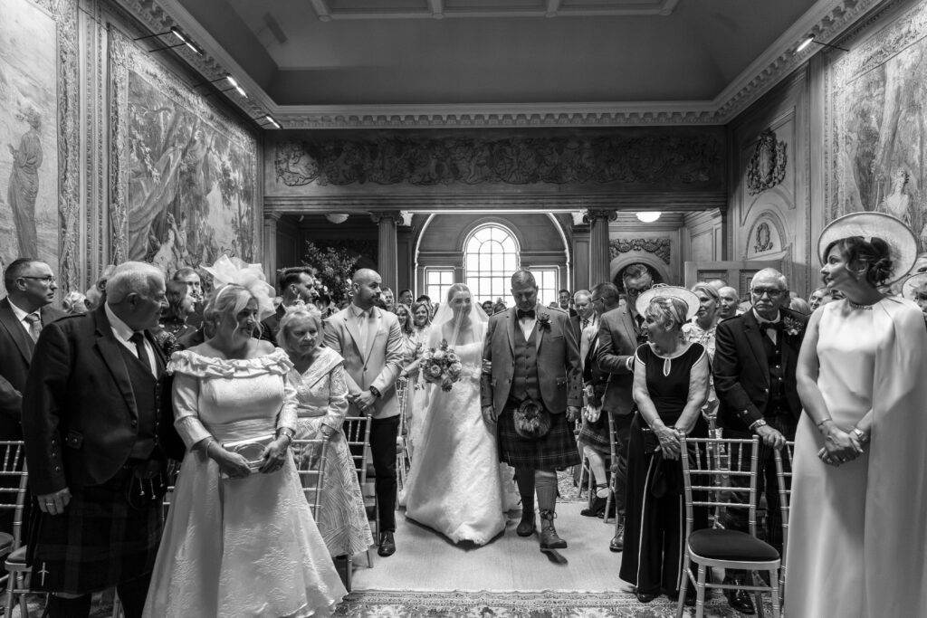 Bride walks down the aisle with her father during a traditional wedding ceremony inside an elegant historic venue, while family and guests stand to watch. Captured in black and white, the bride wears a lace wedding dress and veil, carrying a bouquet as she makes her entrance through a grand room adorned with ornate tapestries and classic architectural details.