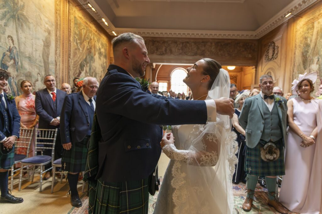 Bride and groom smile at each other during their wedding ceremony, moments after exchanging vows, with the groom wearing a traditional Scottish kilt and the bride in a long-sleeved lace wedding dress and veil. Family and guests look on inside an elegant historic venue with ornate tapestries and warm wooden interiors.
