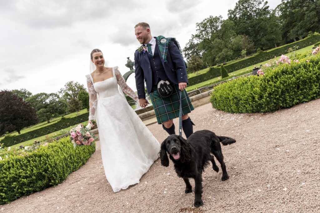Bride and groom walk hand in hand through a formal garden with their black dog leading the way on a leash. The bride wears a long-sleeved lace wedding dress and carries a pink and white bouquet, while the groom wears a traditional Scottish kilt, creating a joyful outdoor wedding portrait against a backdrop of manicured gardens and dramatic skies.