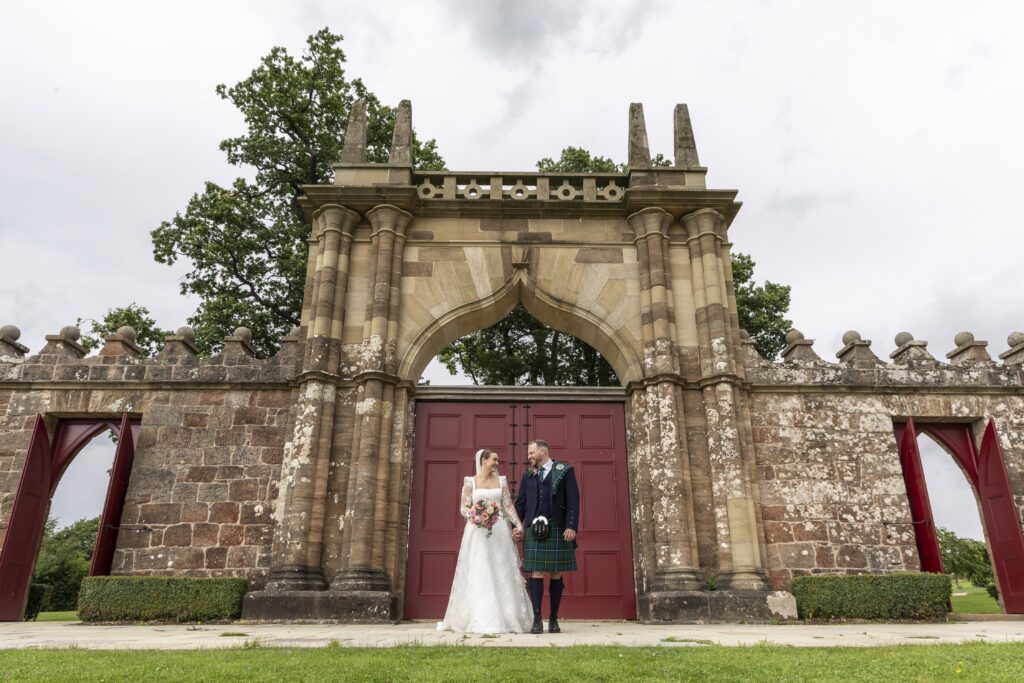 Bride and groom stand hand in hand beneath a grand historic stone archway with striking red doors, smiling at each other during their wedding portraits. The bride wears a lace wedding dress and holds a pink and white bouquet, while the groom wears a traditional Scottish kilt, with the elegant estate architecture and landscaped grounds creating a timeless backdrop.