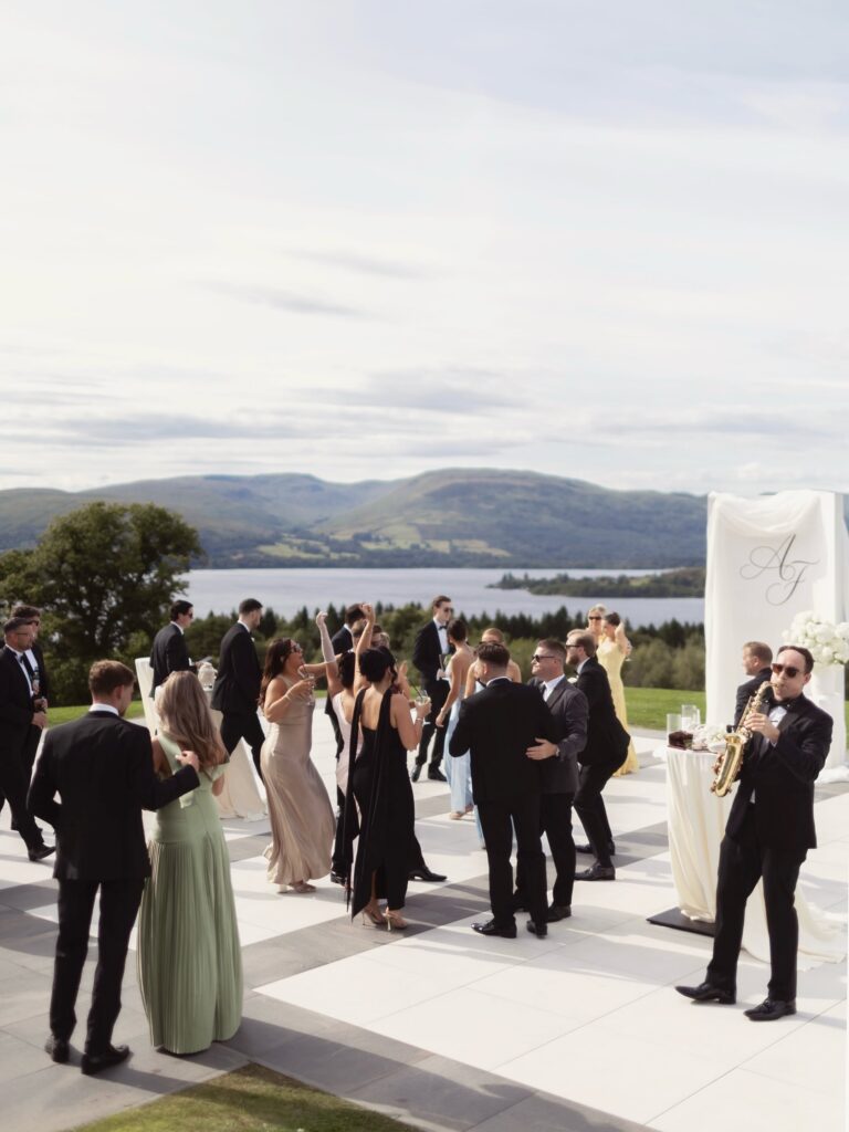 Wedding guests enjoying an outdoor celebration beside a scenic lake and mountains, with a saxophonist performing near a white floral wedding backdrop.