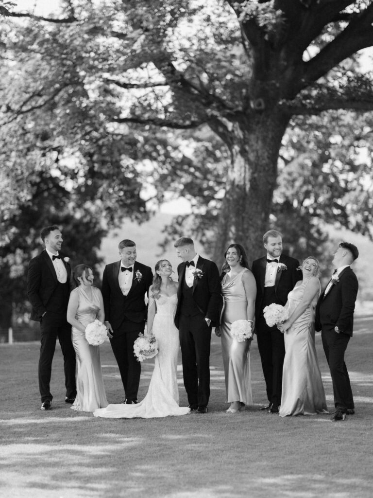 Black-and-white wedding party portrait of the bride and groom standing with their bridesmaids and groomsmen on a lawn beneath a large tree, with everyone smiling and laughing.