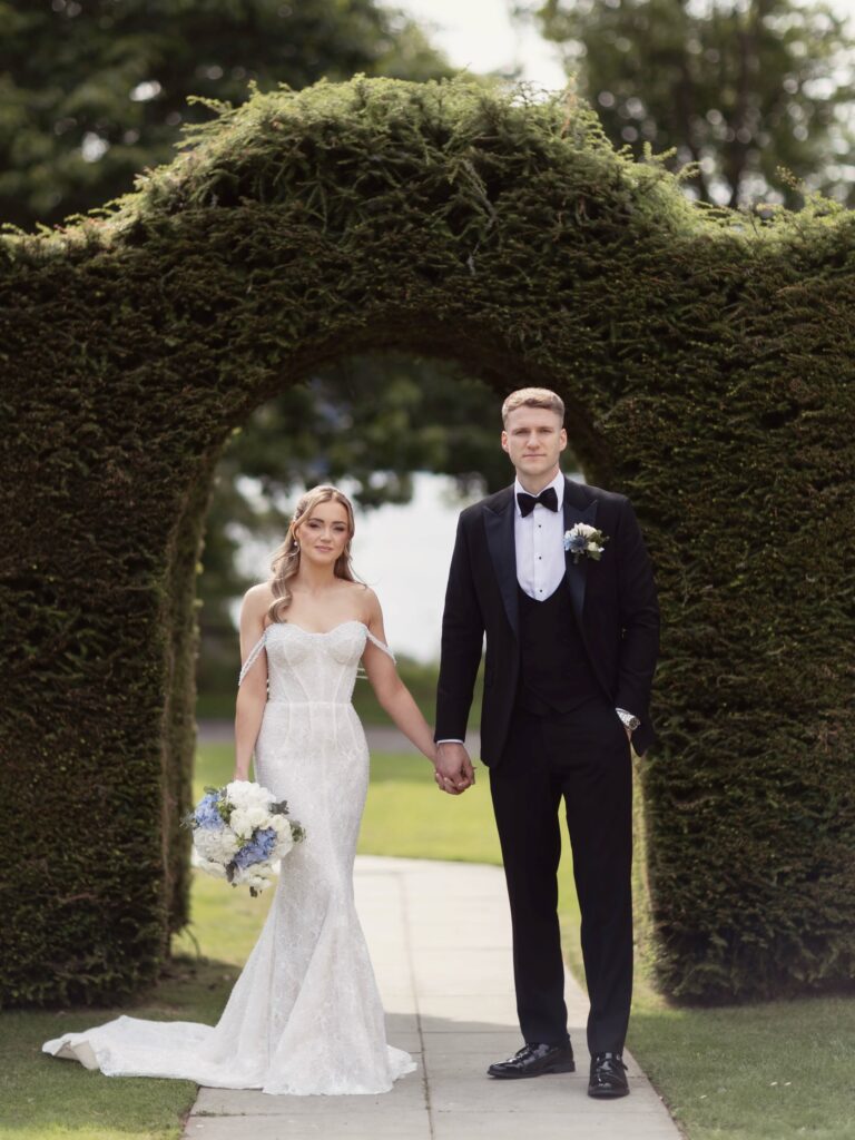 Bride and groom holding hands and posing beneath a lush green garden arch, with the bride carrying a white and blue bouquet.