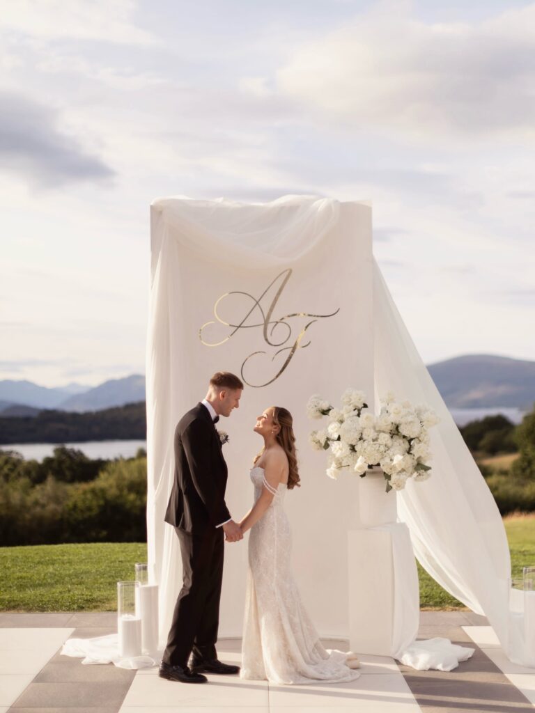 Bride and groom holding hands beneath a white draped wedding arch overlooking a scenic lake and mountains, with white floral arrangements beside them.
