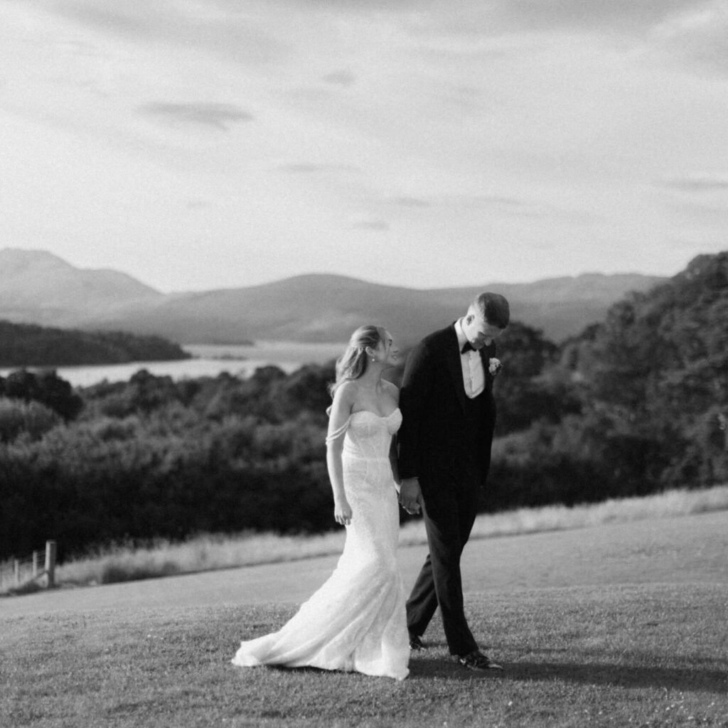 Black-and-white wedding portrait of a bride and groom walking hand in hand across a grassy landscape, with a scenic lake and mountains in the background.