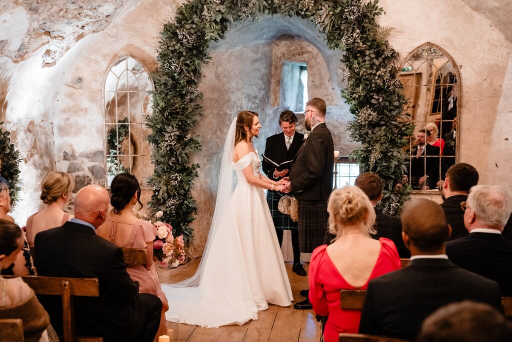 Bride and groom exchange vows beneath a lush floral arch during their Neidpath Castle wedding ceremony