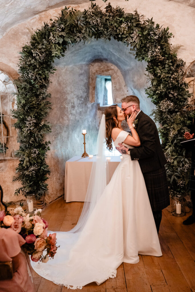 Bride and groom share a kiss beneath a lush floral arch during their Scottish castle wedding ceremony