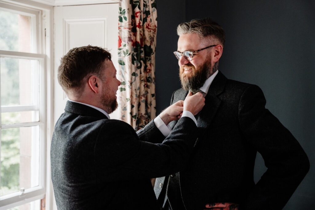 Groom adjusts his tie with help from a groomsman during wedding preparations at a Scottish castle