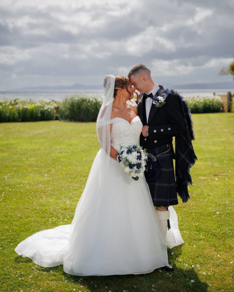 Bride and groom embracing outdoors by the seaside, with the bride holding a white and blue wedding bouquet and the groom wearing a traditional Scottish kilt.