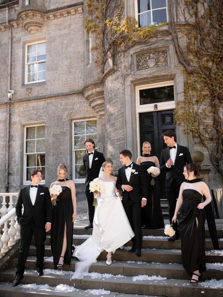 Bride and groom posing with their wedding party on the steps of a historic stone building, surrounded by elegant architecture and white wedding confetti.