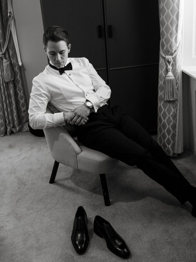 Black-and-white portrait of a groom in a white shirt and bow tie sitting in a chair, with polished black dress shoes on the floor nearby.