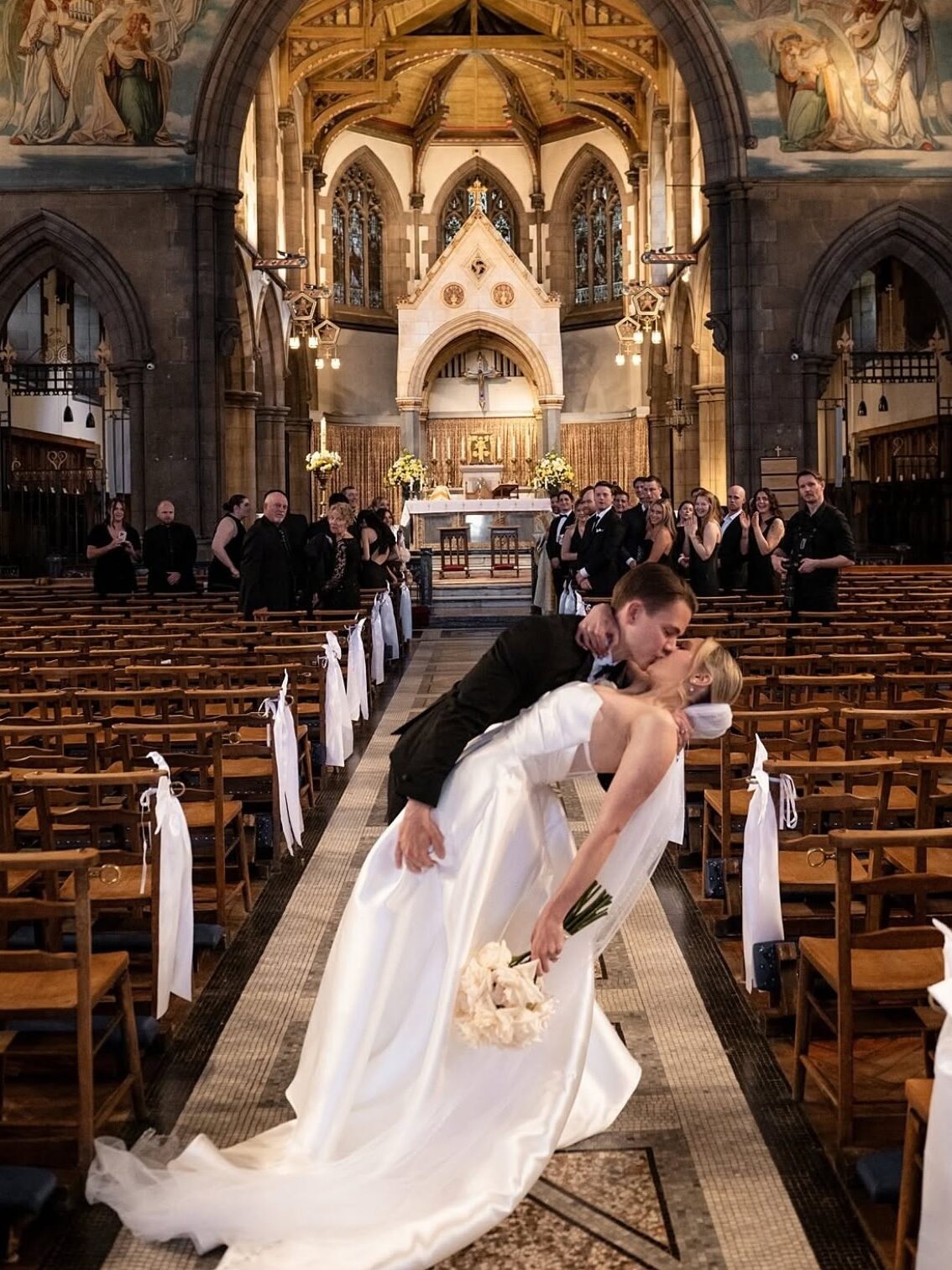 Bride and groom sharing a kiss in the aisle of a grand church, surrounded by wedding guests and ornate religious artwork.