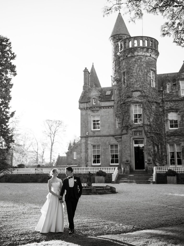 Black-and-white wedding portrait of a bride and groom walking together in front of a historic stone castle with a turret.