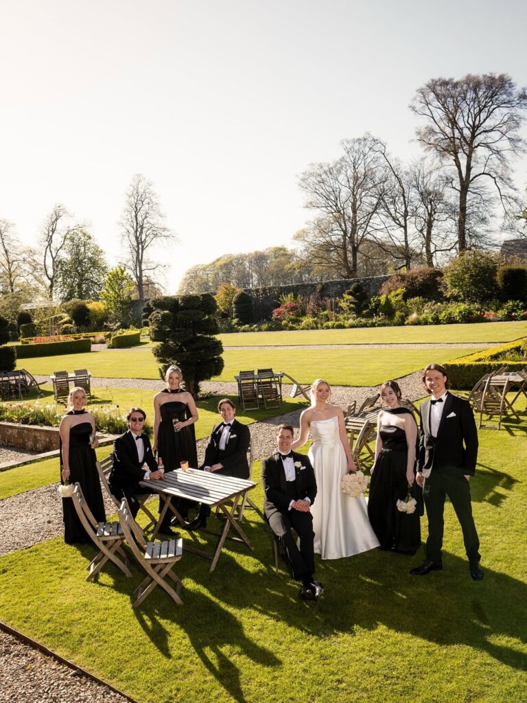 Bride and groom posing with their wedding party in a formal garden, surrounded by manicured lawns, topiary trees, and outdoor seating.