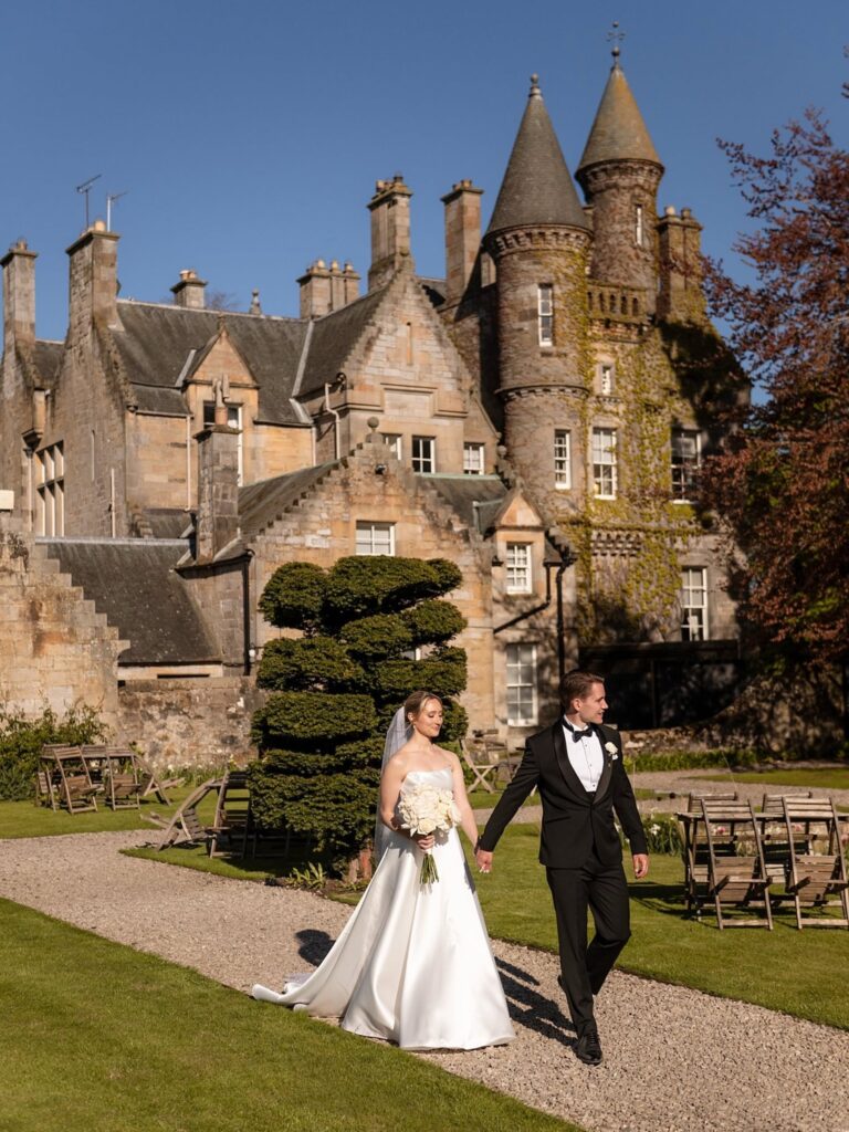 Bride and groom walking hand in hand through the grounds of a historic stone castle on a sunny wedding day.