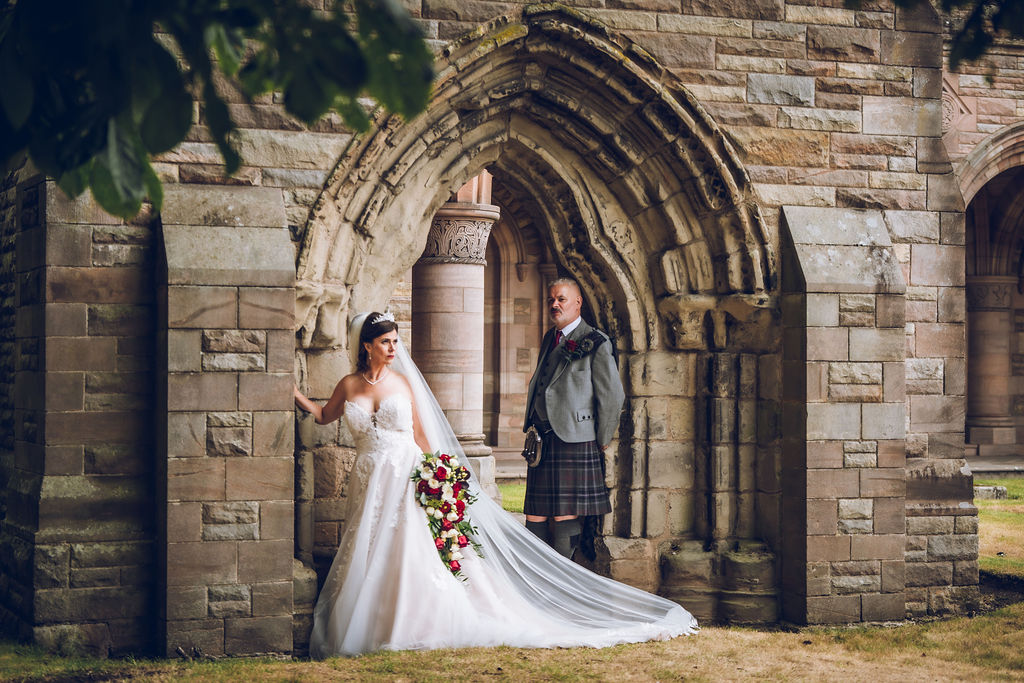 Bride in a white wedding gown holding a cascading bouquet stands beneath a historic stone archway while the groom, dressed in a traditional Scottish kilt, looks on in a romantic outdoor wedding portrait.